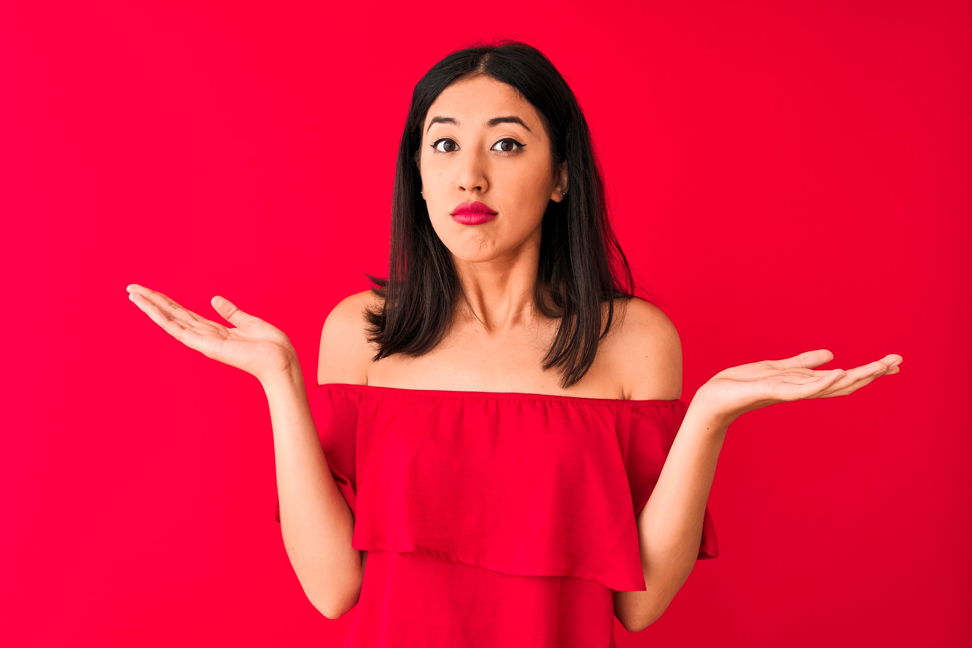 A woman with long dark hair is wearing a red off-the-shoulder top against a red background. She has an expression of confusion or uncertainty, with her arms outstretched and palms facing up.