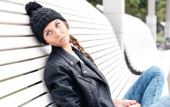 Woman in a black knit hat and leather jacket sitting on a white wooden bench. She has a serious expression, with her head tilted slightly back, and her long hair is braided. There's a blurry background of greenery and concrete pillars.