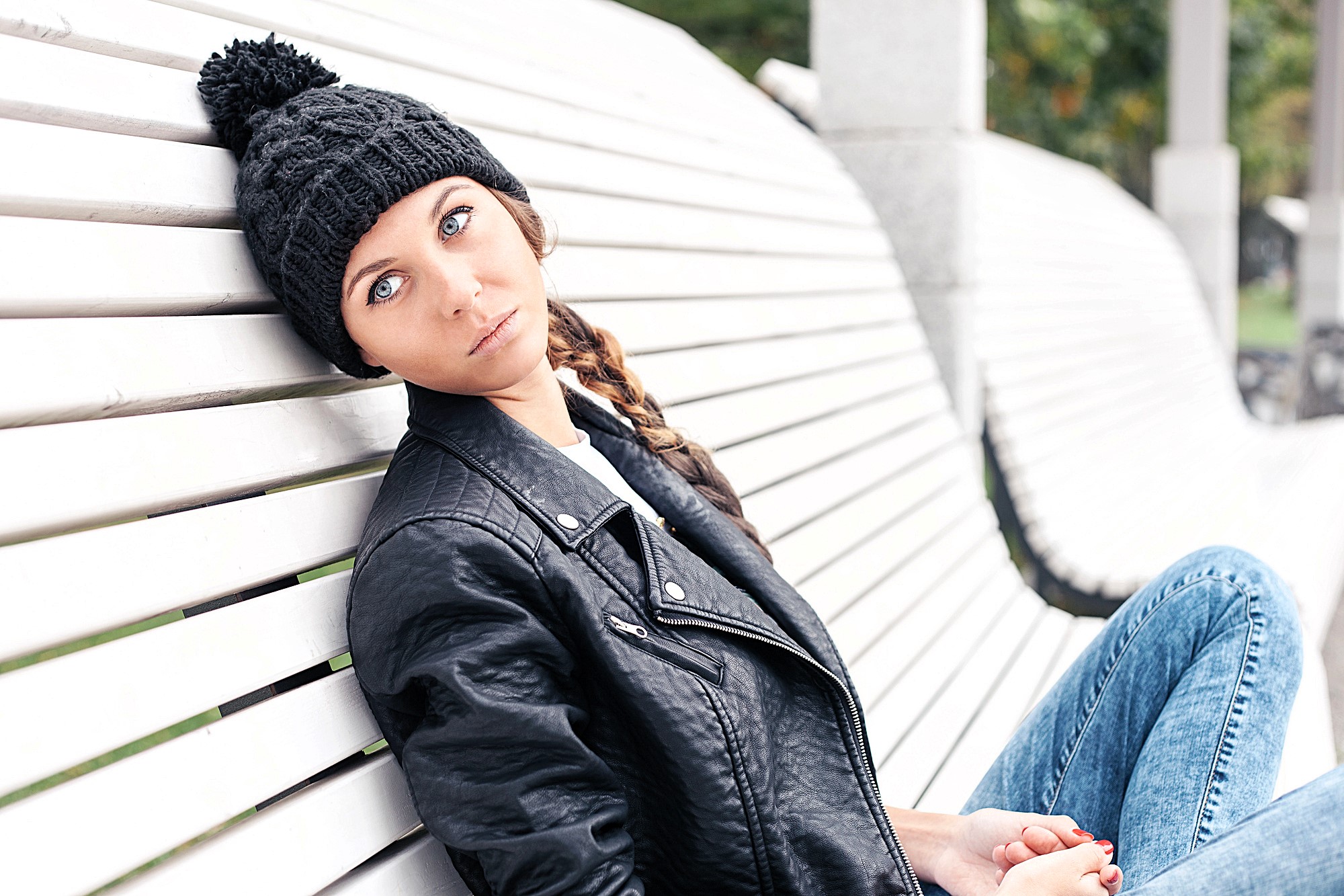 Woman in a black knit hat and leather jacket sitting on a white wooden bench. She has a serious expression, with her head tilted slightly back, and her long hair is braided. There's a blurry background of greenery and concrete pillars.