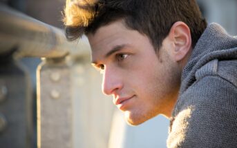 A young man with short, dark hair gazes thoughtfully into the distance. He is wearing a gray knit sweater and stands near a metal railing, with soft sunlight illuminating his face. The background is softly blurred.