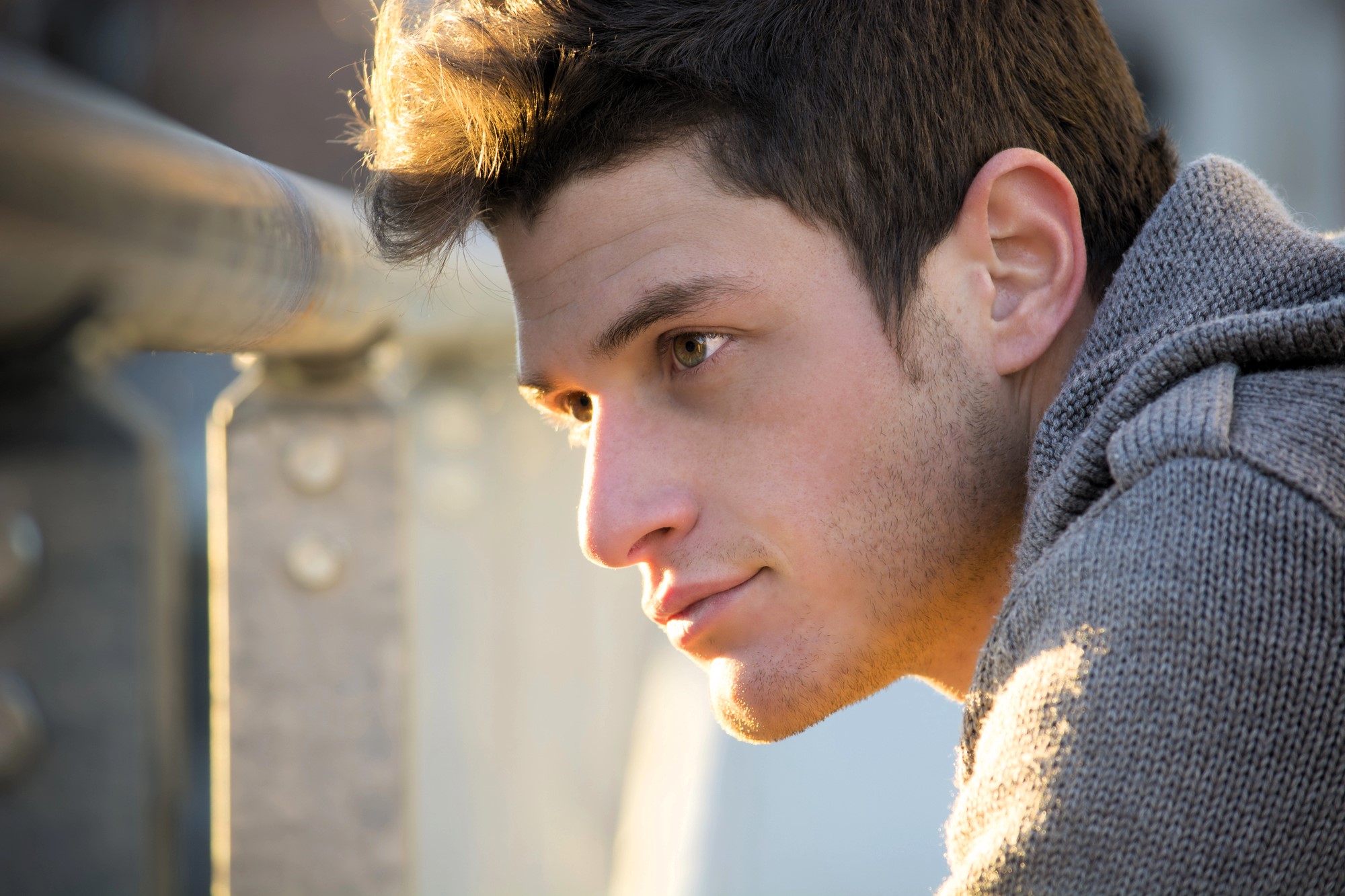 A young man with short, dark hair gazes thoughtfully into the distance. He is wearing a gray knit sweater and stands near a metal railing, with soft sunlight illuminating his face. The background is softly blurred.