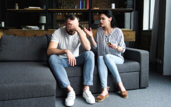 A man and woman sit on a gray couch in a loft-style room. The man covers his face with one hand, appearing distressed. The woman gestures with one hand, holding glasses in the other, as if speaking to him. Shelves and books are visible in the background.