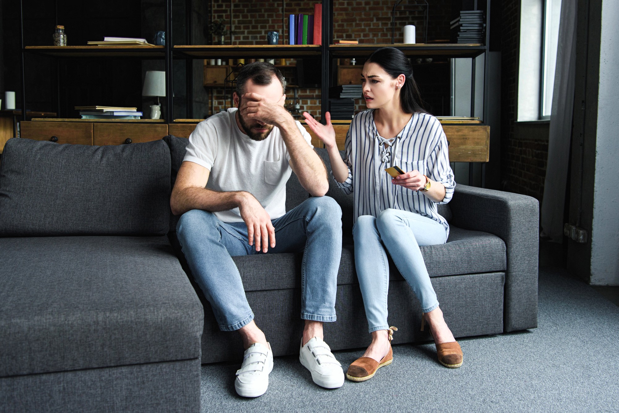 A man and woman sit on a gray couch in a loft-style room. The man covers his face with one hand, appearing distressed. The woman gestures with one hand, holding glasses in the other, as if speaking to him. Shelves and books are visible in the background.