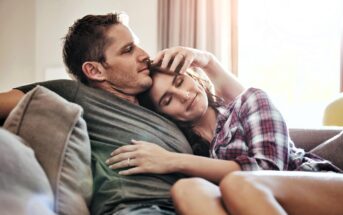 A couple sits closely on a sofa, with the man resting his head against the woman's. The woman, wearing a plaid shirt, has her eyes closed and smiles gently. Sunlight filters through a window, creating a warm and cozy atmosphere.