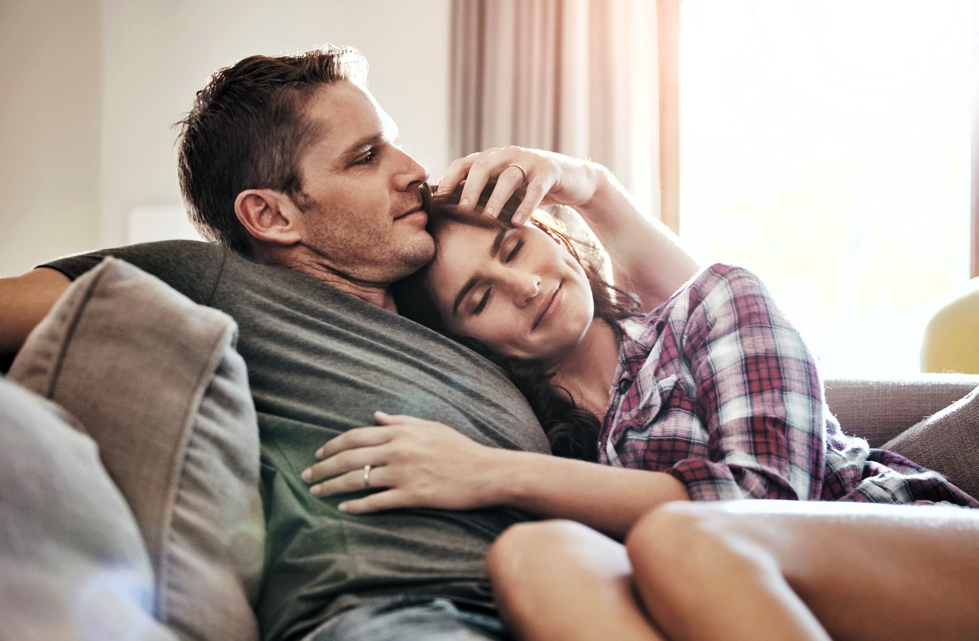 A couple sits closely on a sofa, with the man resting his head against the woman's. The woman, wearing a plaid shirt, has her eyes closed and smiles gently. Sunlight filters through a window, creating a warm and cozy atmosphere.