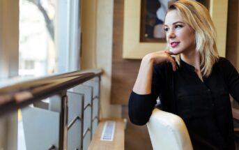 A woman with blonde hair sits indoors, gazing out the window, resting her chin on her hand. She wears a black top and appears thoughtful. The setting includes a framed photo on the wall and soft lighting.