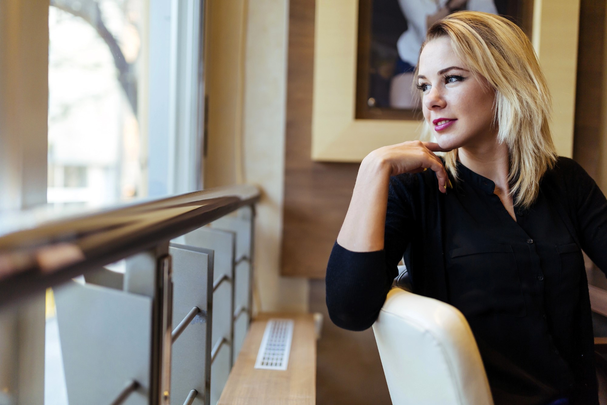 A woman with blonde hair sits indoors, gazing out the window, resting her chin on her hand. She wears a black top and appears thoughtful. The setting includes a framed photo on the wall and soft lighting.