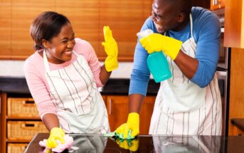 A man and woman in aprons, wearing yellow gloves, enjoy cleaning a kitchen together. They are smiling and playfully interacting while holding cleaning sprays and cloths. The kitchen has wooden cabinets and a black countertop.