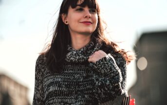 A woman with long brown hair and a fringe is walking outside, wearing a knitted sweater. She is holding a striped shopping bag and smiling, with blurred buildings in the background. The sunlight casts a natural glow on her face.