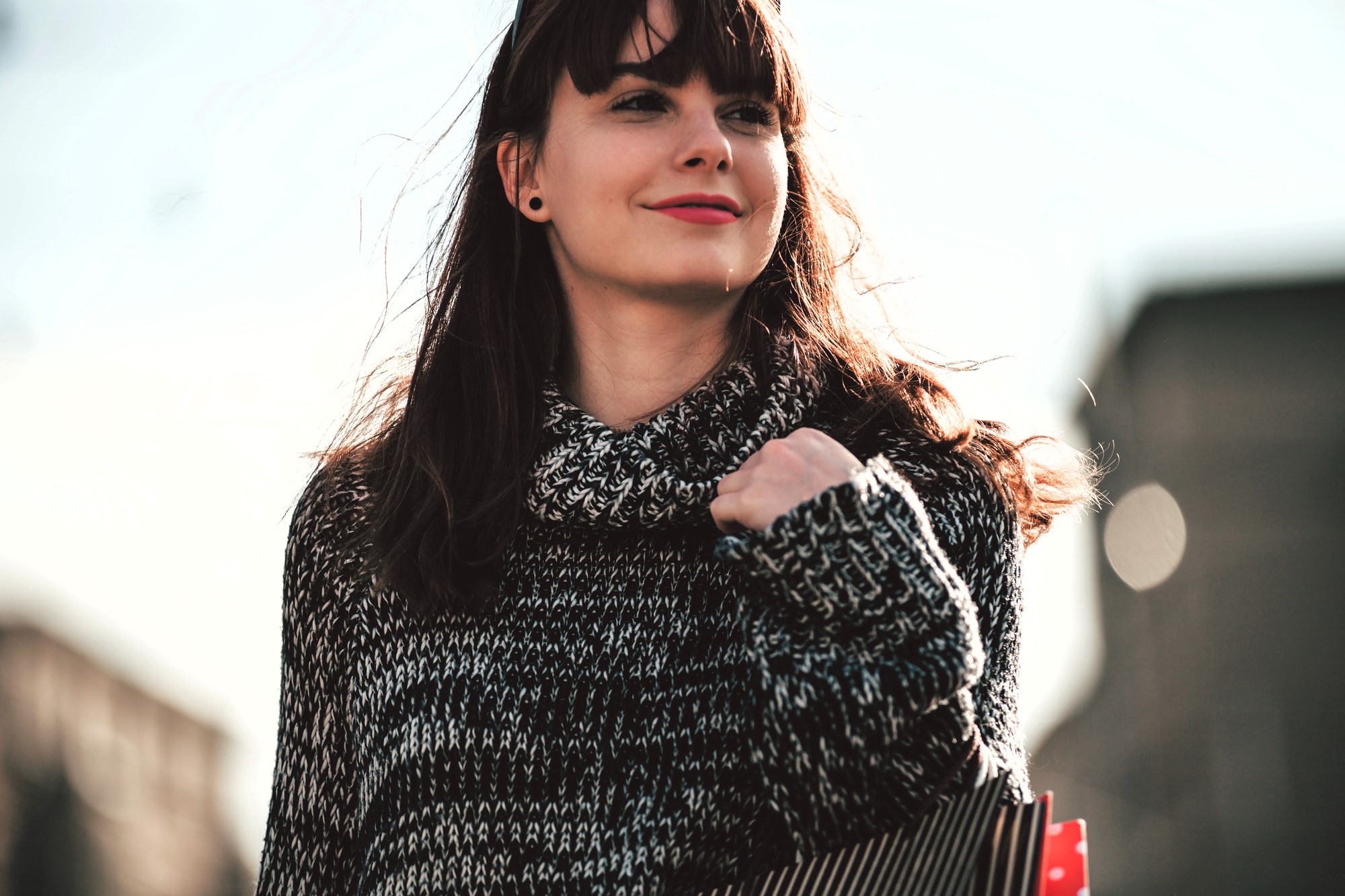 A woman with long brown hair and a fringe is walking outside, wearing a knitted sweater. She is holding a striped shopping bag and smiling, with blurred buildings in the background. The sunlight casts a natural glow on her face.