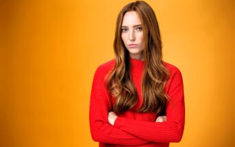 A long-haired person with a serious expression stands with arms crossed, wearing a bright red sweater. The background is a solid orange.