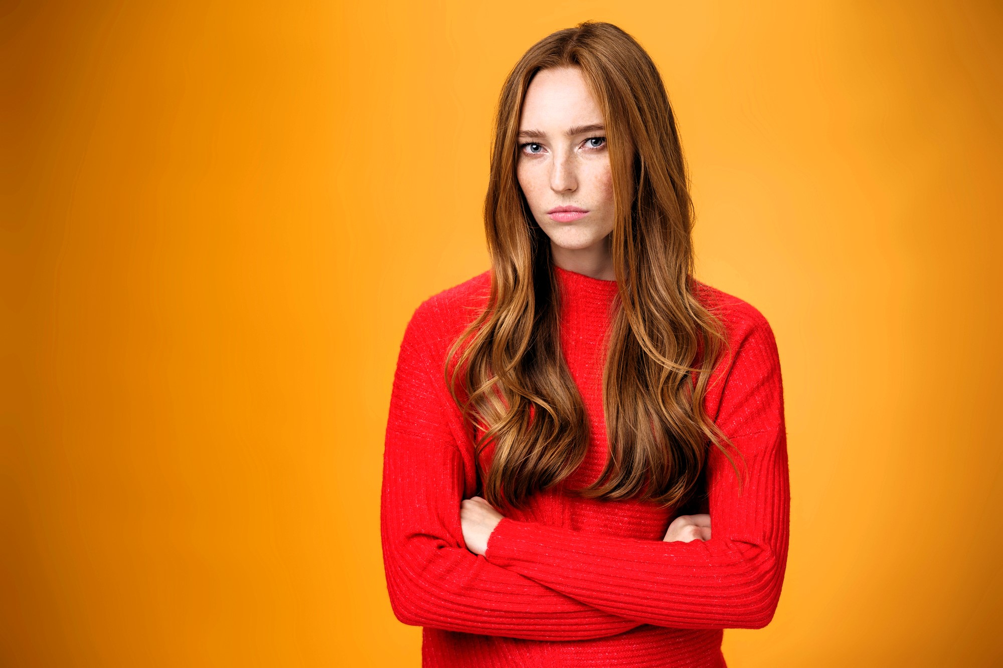 A long-haired person with a serious expression stands with arms crossed, wearing a bright red sweater. The background is a solid orange.