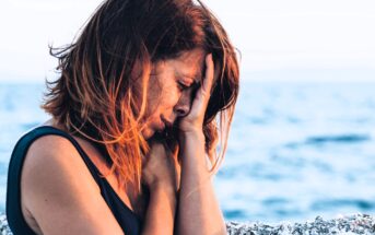 A woman with red hair in a dark top sits by the ocean, her hand covering her face as if in distress or deep thought. The sea is visible in the blurred background, suggesting a contemplative or emotional moment.