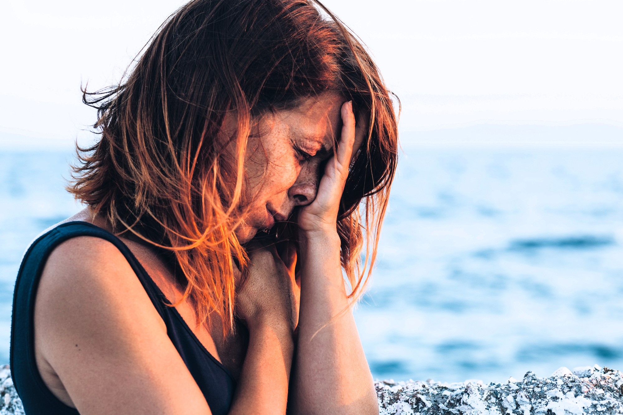 A woman with red hair in a dark top sits by the ocean, her hand covering her face as if in distress or deep thought. The sea is visible in the blurred background, suggesting a contemplative or emotional moment.