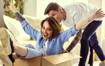 A woman sits playfully in a cardboard box with her arms spread wide, smiling and wearing a blue shirt and jeans. A man with tattoos in a white shirt playfully pushes the box. They are indoors with light streaming through a window.