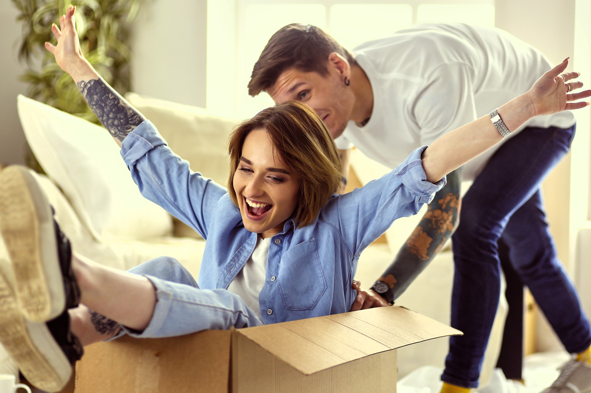 A woman sits playfully in a cardboard box with her arms spread wide, smiling and wearing a blue shirt and jeans. A man with tattoos in a white shirt playfully pushes the box. They are indoors with light streaming through a window.