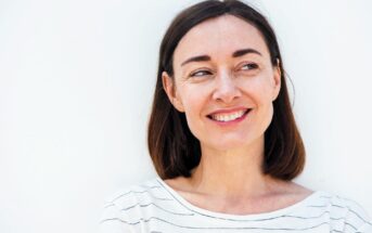 A woman with shoulder-length brown hair and wearing a striped shirt smiles while looking to the side. The background is a plain white wall.