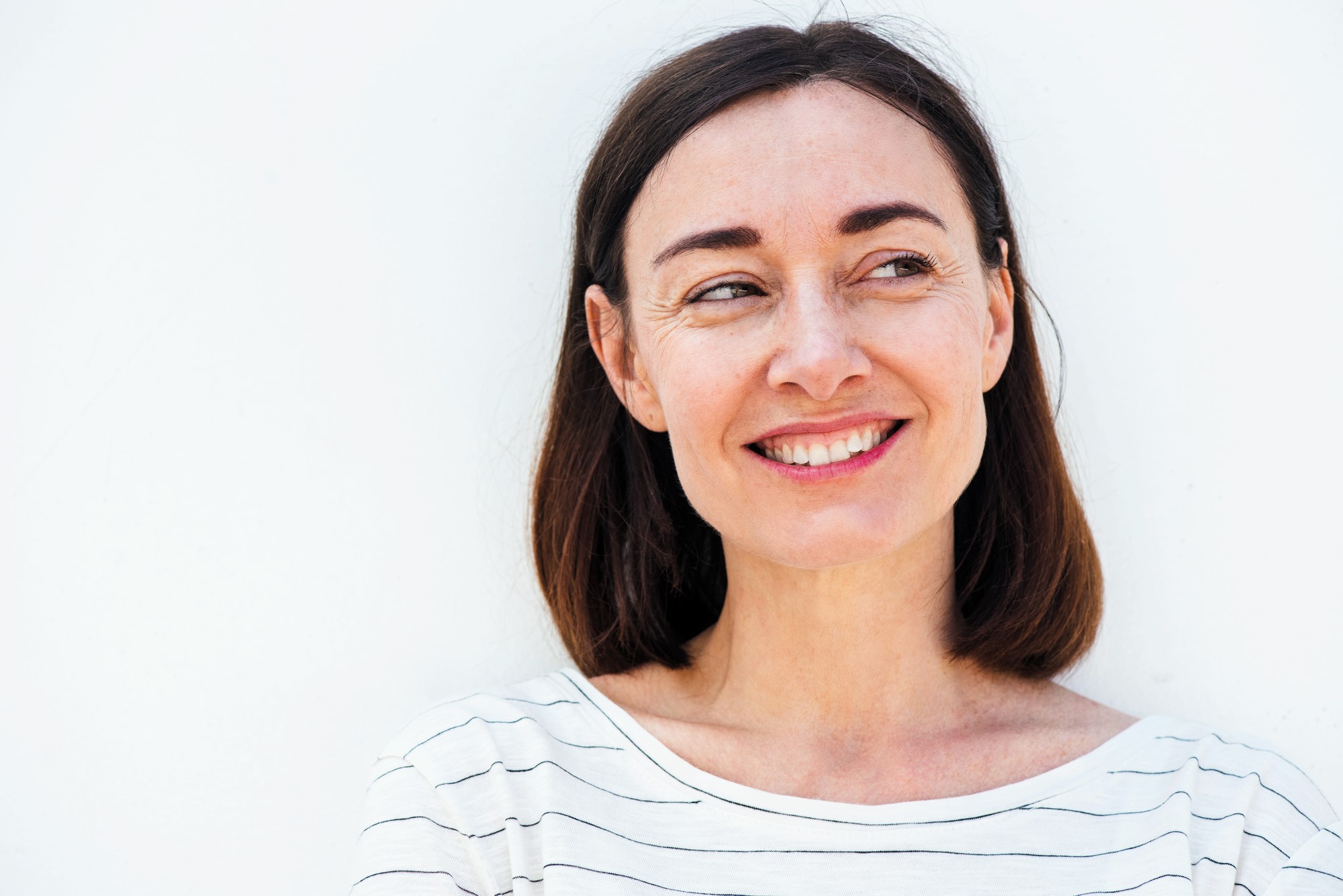 A woman with shoulder-length brown hair and wearing a striped shirt smiles while looking to the side. The background is a plain white wall.