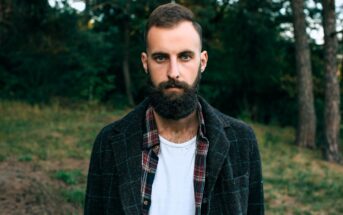 A bearded man stands outdoors in a forested area. He is wearing a dark checkered jacket over a red plaid shirt and a white t-shirt. Tall trees are visible in the background.