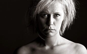 Black and white portrait of a person with short, tousled hair, looking directly at the camera. The lighting is dramatic, highlighting their facial features and creating shadows on the right side. The background is dark.