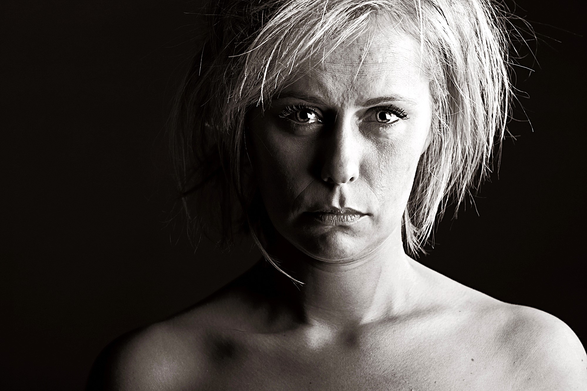 Black and white portrait of a person with short, tousled hair, looking directly at the camera. The lighting is dramatic, highlighting their facial features and creating shadows on the right side. The background is dark.