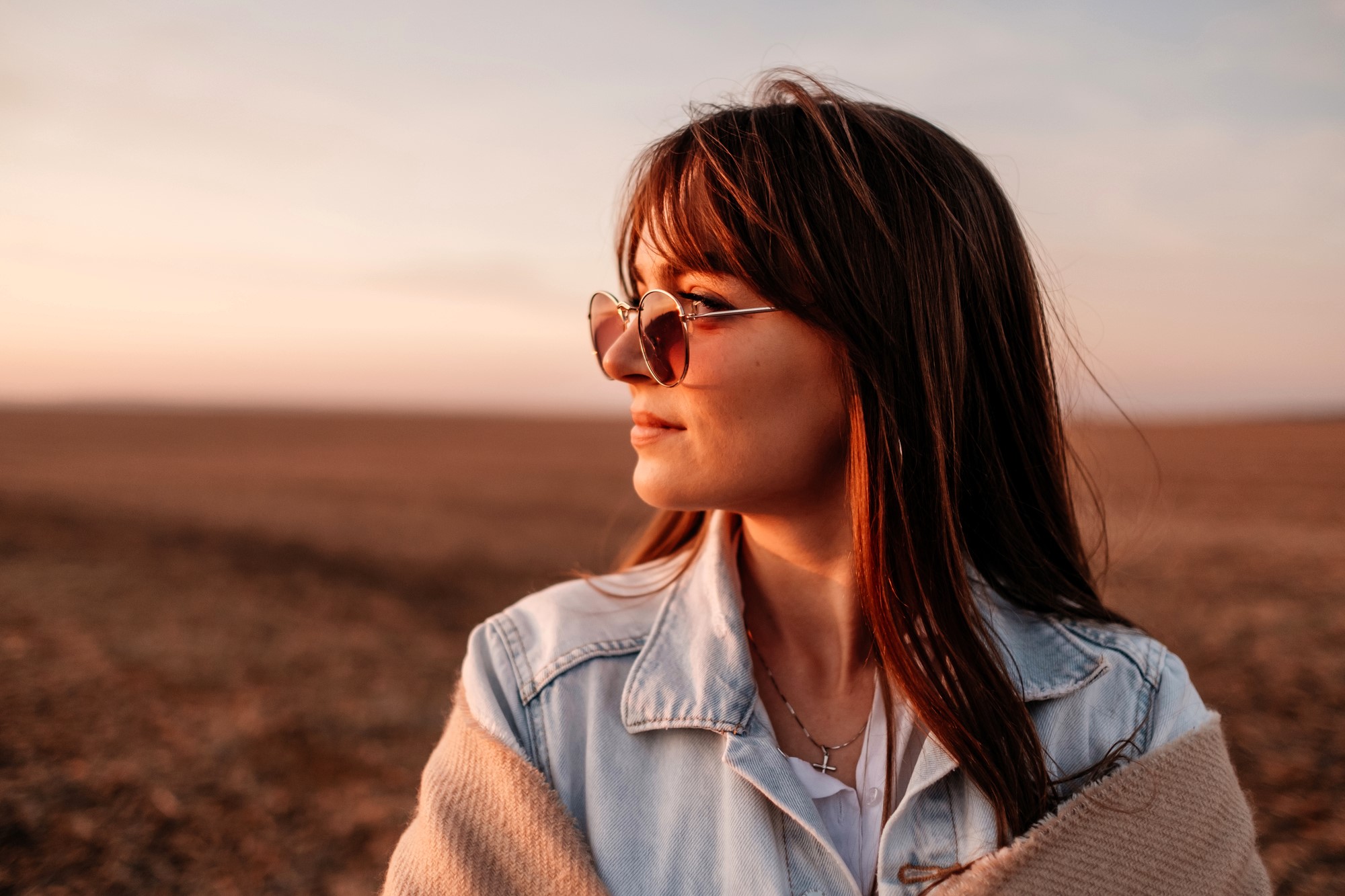 A woman with long hair and sunglasses stands in an open field at sunset. She wears a denim jacket and has a cross necklace. The sky is softly lit with gentle hues of orange and pink.