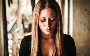 A woman with long brown hair is looking down with her eyes closed. She is wearing a black top and is in a softly lit, blurred background setting, suggesting an indoor or sheltered location.