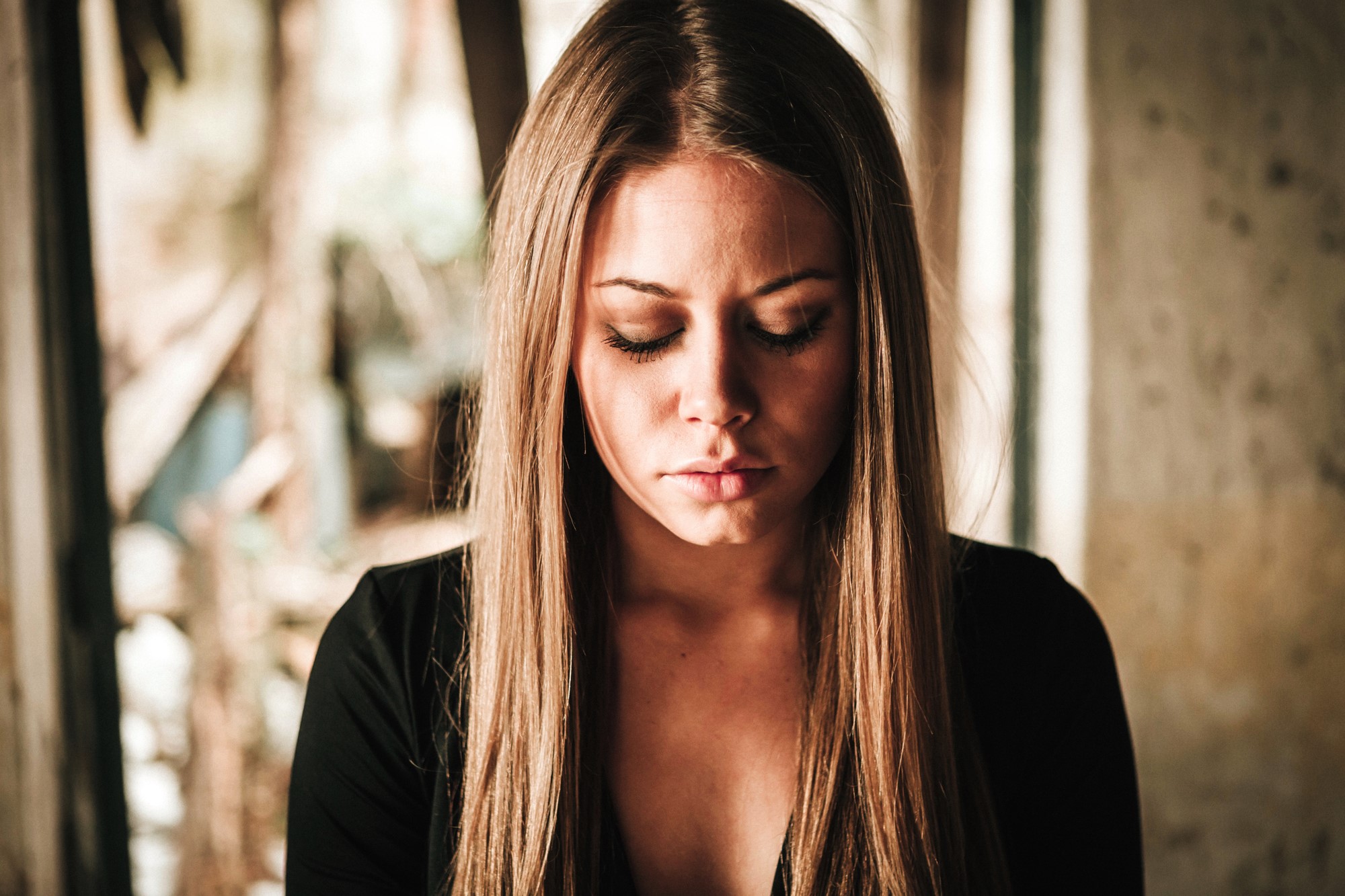 A woman with long brown hair is looking down with her eyes closed. She is wearing a black top and is in a softly lit, blurred background setting, suggesting an indoor or sheltered location.