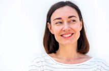 A woman with shoulder-length brown hair smiles while looking to the side. She is wearing a white and black striped shirt, standing against a plain white background.