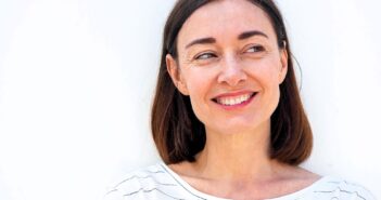 A woman with shoulder-length brown hair smiles while looking to the side. She is wearing a white and black striped shirt, standing against a plain white background.