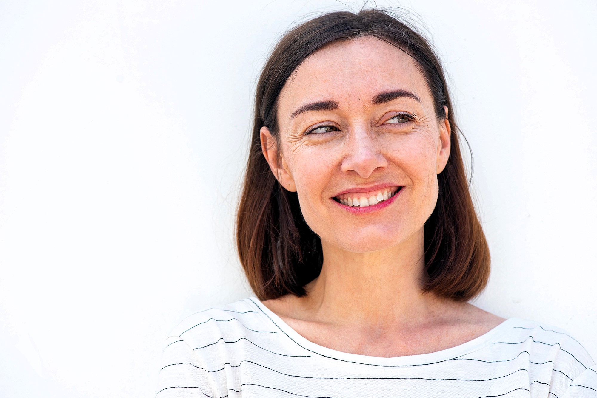 A woman with shoulder-length brown hair smiles while looking to the side. She is wearing a white and black striped shirt, standing against a plain white background.