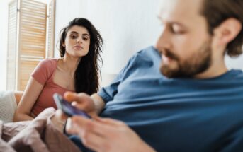 A woman with long dark hair in a pink shirt looks towards a man with a beard in a blue shirt, who is focused on a smartphone. They are sitting on a bed, with wooden blinds partially visible in the background.