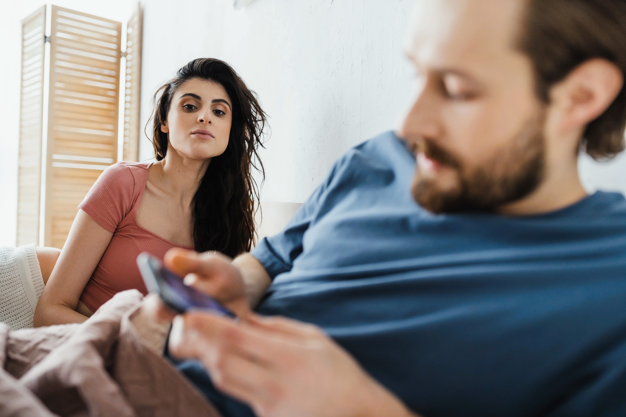 A woman with long dark hair in a pink shirt looks towards a man with a beard in a blue shirt, who is focused on a smartphone. They are sitting on a bed, with wooden blinds partially visible in the background.