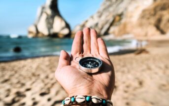 An outstretched hand holds a compass on a sandy beach with turquoise water in the background. Rocky cliffs are visible under a clear blue sky. The person is wearing a seashell bracelet.