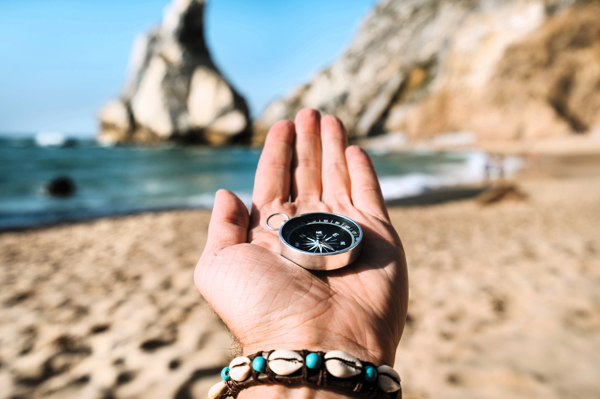 An outstretched hand holds a compass on a sandy beach with turquoise water in the background. Rocky cliffs are visible under a clear blue sky. The person is wearing a seashell bracelet.