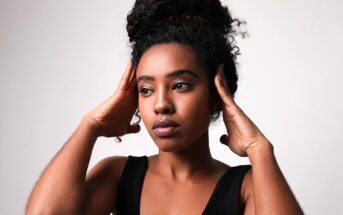 A person with curly hair tied up in a bun rests their hands on the sides of their head, looking to the left. They wear a black sleeveless top and stand against a neutral background, displaying a thoughtful expression.