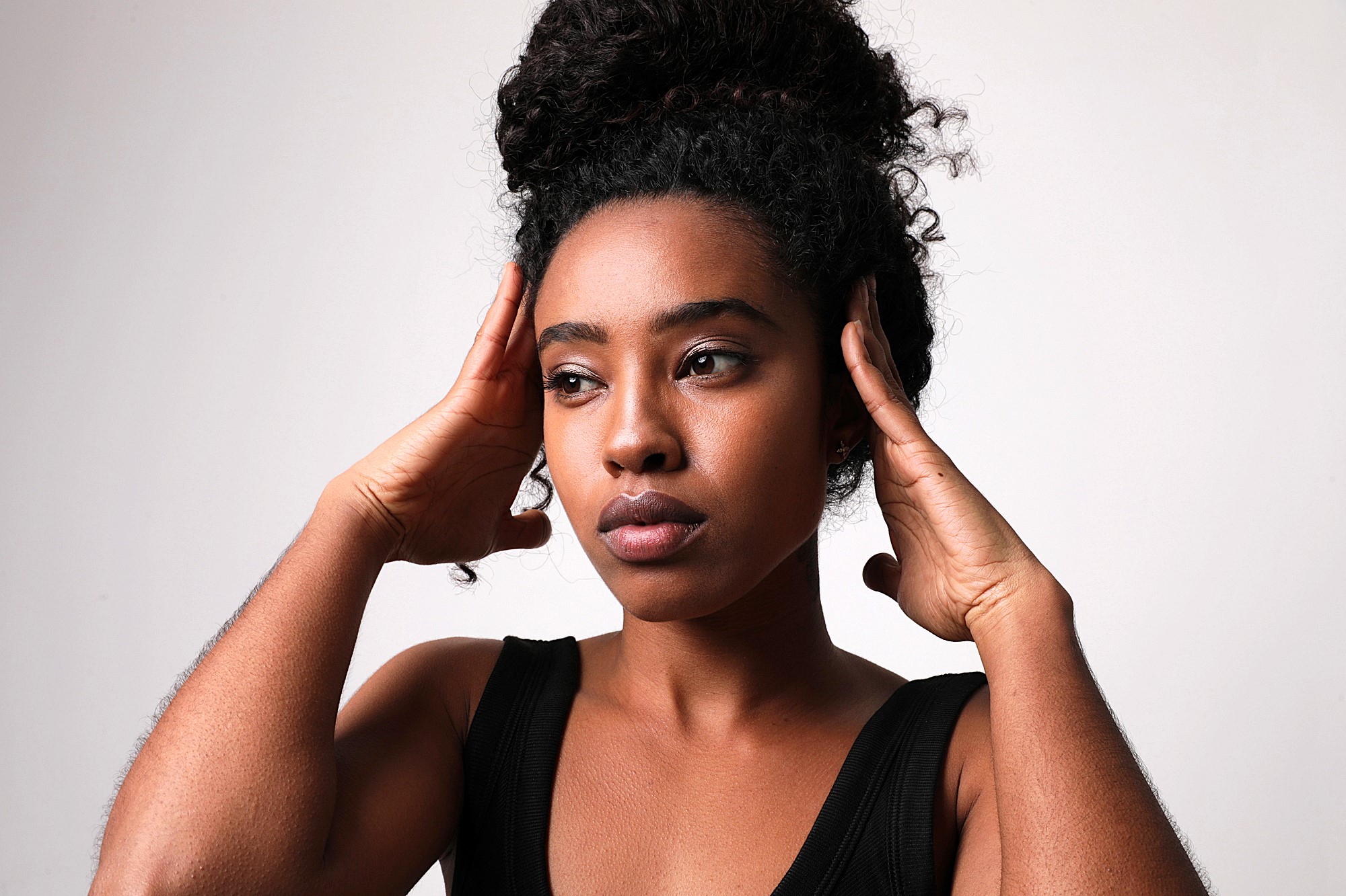 A person with curly hair tied up in a bun rests their hands on the sides of their head, looking to the left. They wear a black sleeveless top and stand against a neutral background, displaying a thoughtful expression.