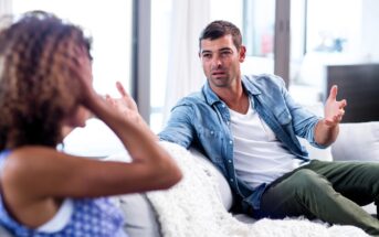 A man in a denim shirt and a woman in a blue patterned top are having a conversation on a sofa. The man gestures with his hands while speaking, and the woman covers her face with her hand. A white blanket is draped over the sofa.