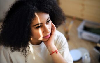 A woman with curly hair and a thoughtful expression rests her chin on her hand. She is wearing a white shirt and a red bracelet. Sunlight casts shadows on her face. The background is softly blurred, with art supplies visible.