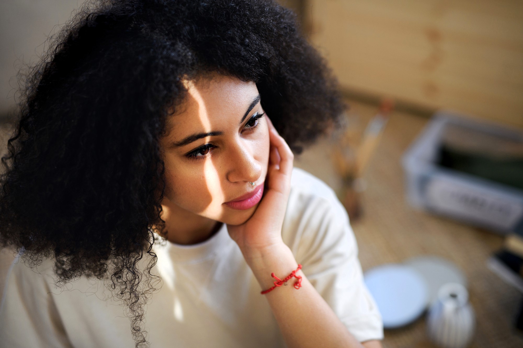 A woman with curly hair and a thoughtful expression rests her chin on her hand. She is wearing a white shirt and a red bracelet. Sunlight casts shadows on her face. The background is softly blurred, with art supplies visible.