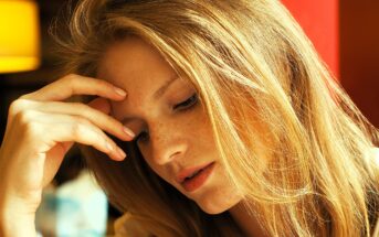 Woman with long, light brown hair and freckles gently touches her forehead with her hand while looking down. She appears thoughtful or pensive. A soft, warm lighting creates a calm ambiance.