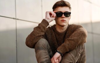 A person with a brown sweater and camouflage pants sits against a wall, partially lifting black sunglasses. The setting is outdoors, with soft lighting, conveying a relaxed and stylish vibe.