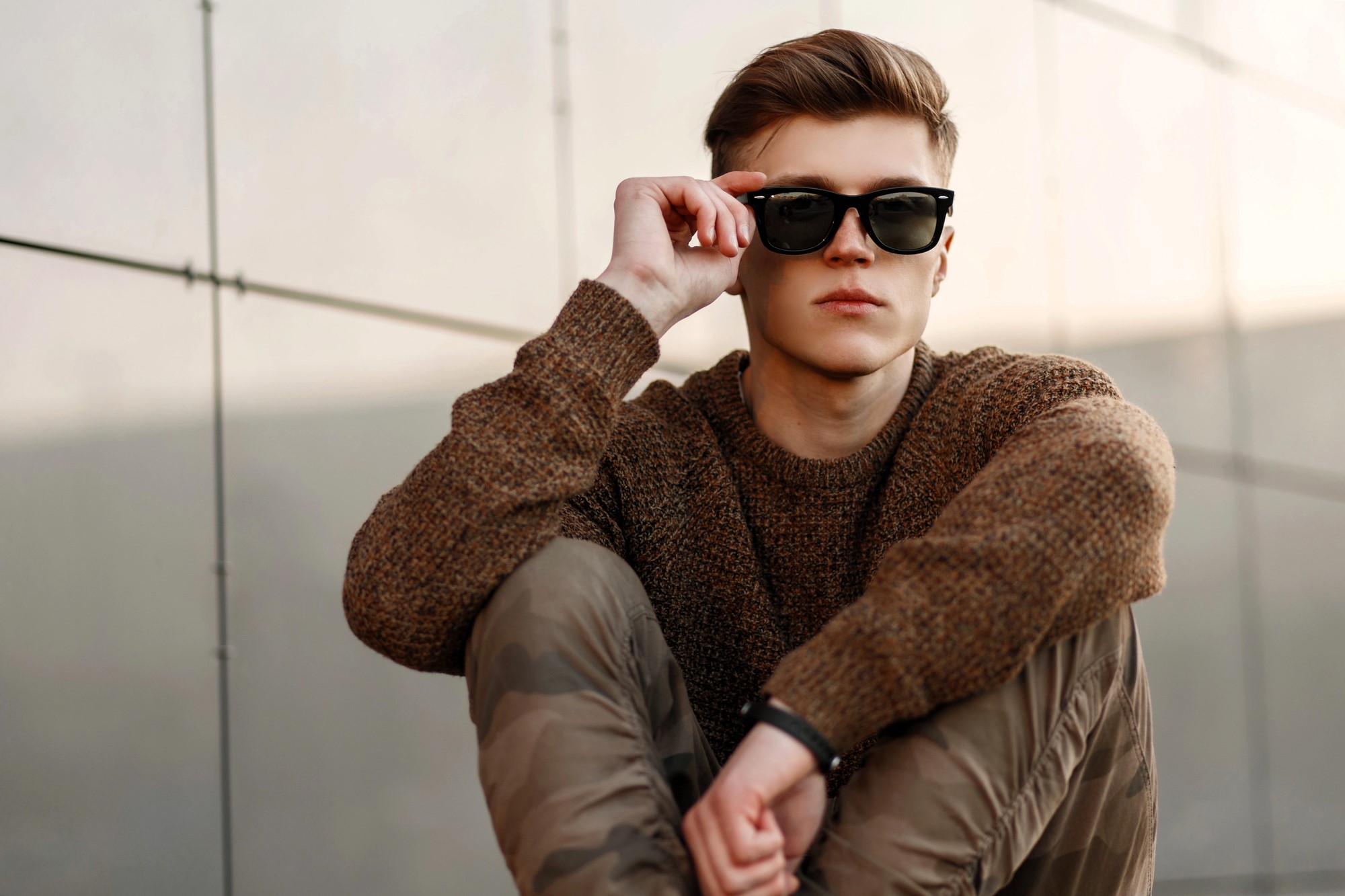 A person with a brown sweater and camouflage pants sits against a wall, partially lifting black sunglasses. The setting is outdoors, with soft lighting, conveying a relaxed and stylish vibe.