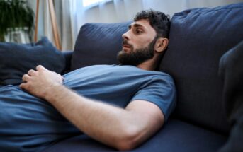 A person with a beard is lying on a blue couch, wearing a blue T-shirt. They have their hands resting on their abdomen and are looking pensive. The background includes a bright window and blurred greenery.