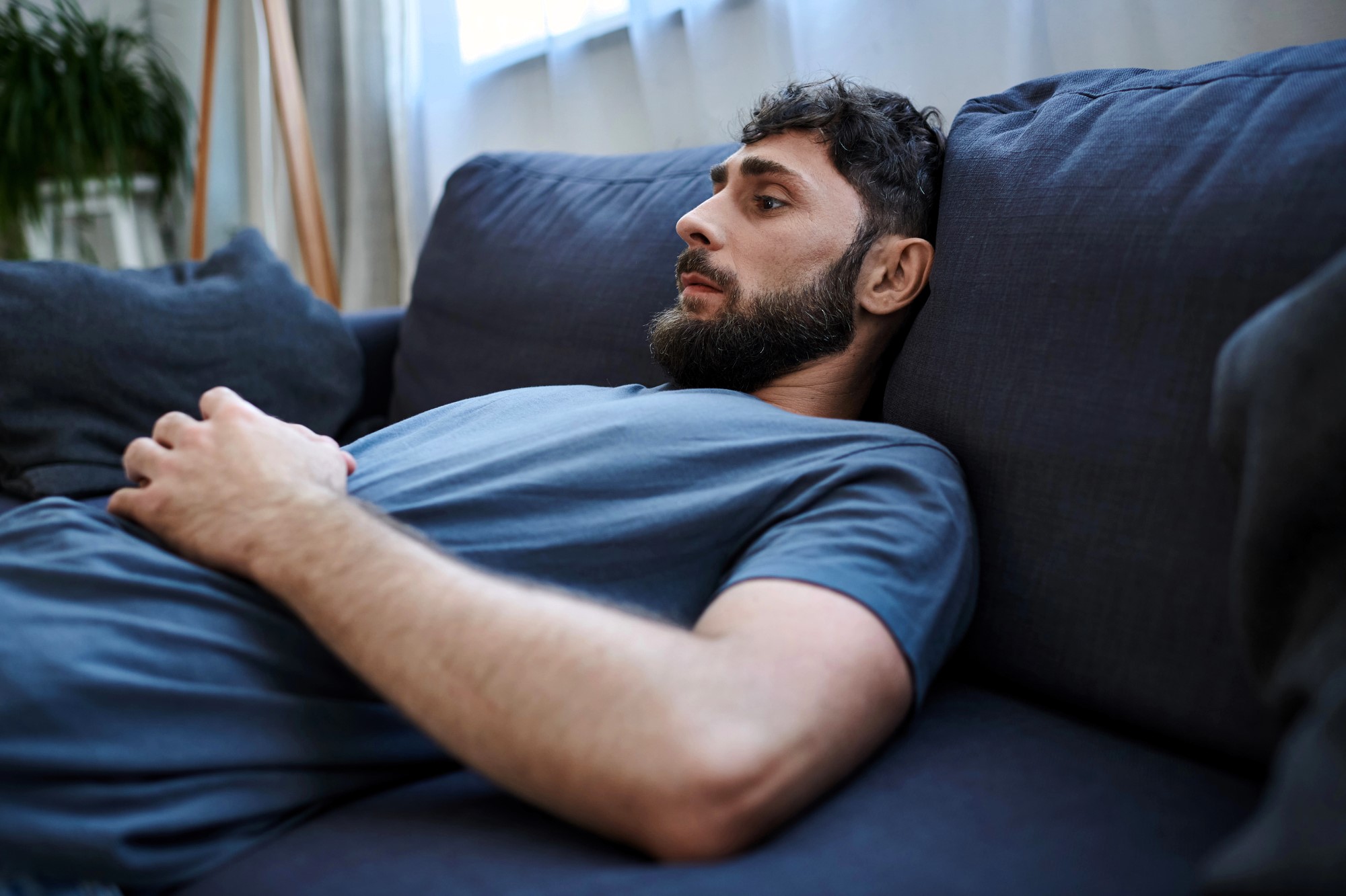 A person with a beard is lying on a blue couch, wearing a blue T-shirt. They have their hands resting on their abdomen and are looking pensive. The background includes a bright window and blurred greenery.