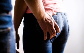 A person wearing jeans is gently touching the back pocket of another person, also in jeans. The second person is facing away, wearing a pink shirt. The scene is well-lit with a neutral background.