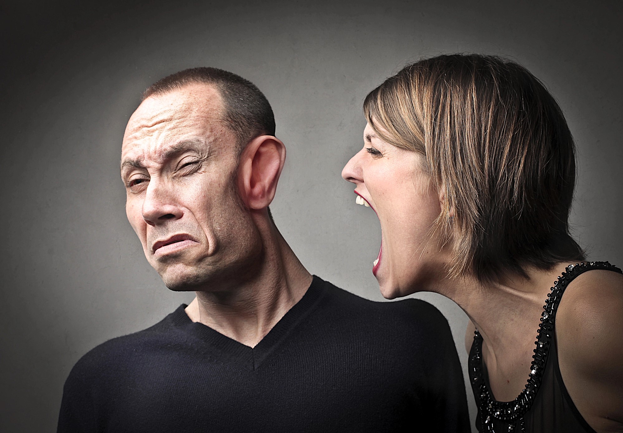 A woman is yelling at a man with exaggeratedly large ears. The man winces and turns away, looking uncomfortable. Both are in casual attire, set against a neutral gray background.