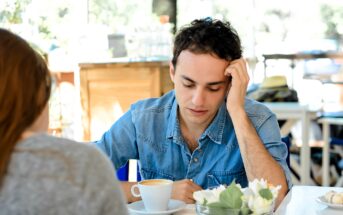 A man in a denim shirt appears thoughtful while sitting at a café table with a cup of coffee. Opposite him, part of another person is visible. The table is decorated with a small vase of white flowers. The background is softly blurred.