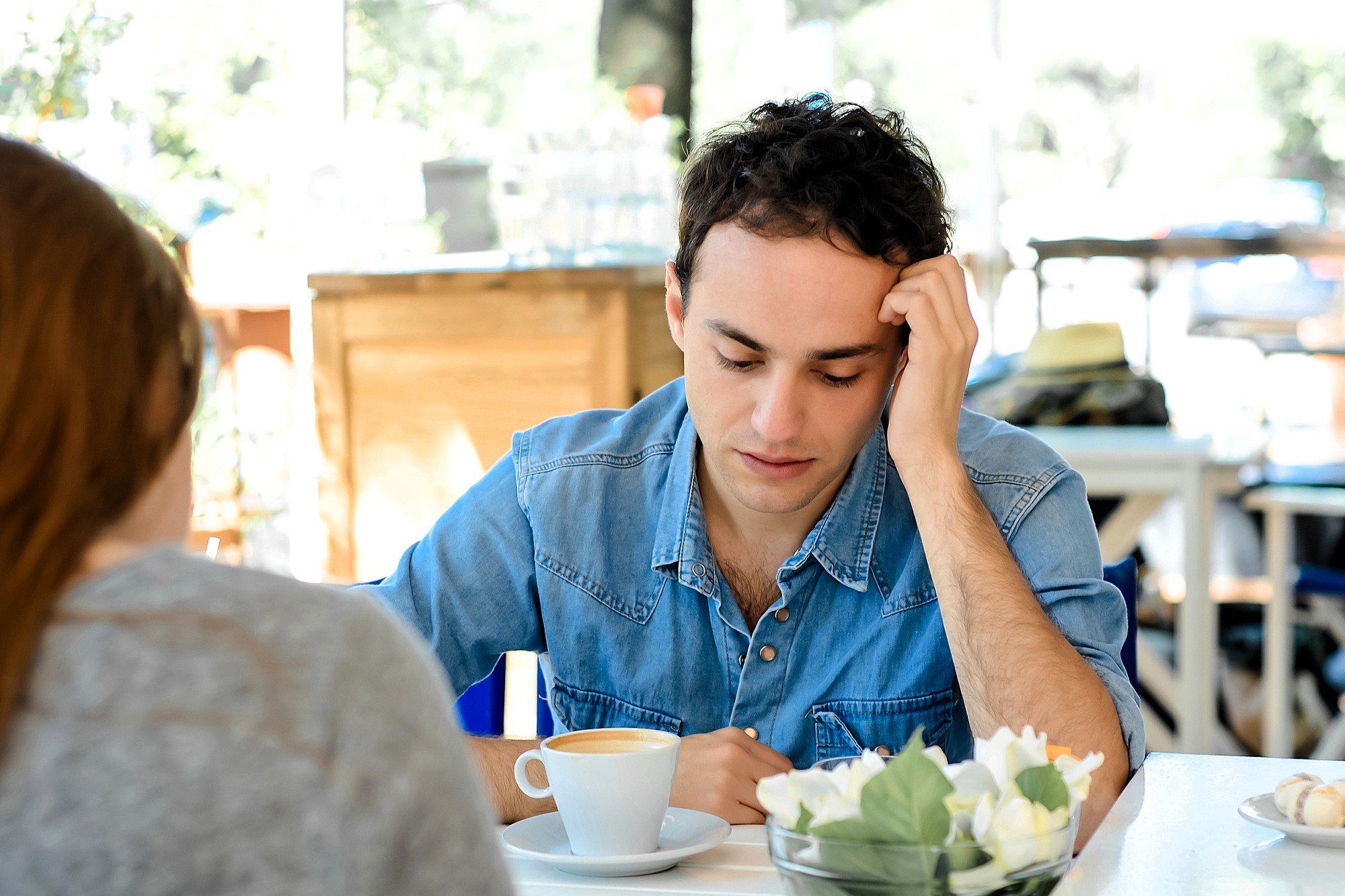 A man in a denim shirt appears thoughtful while sitting at a café table with a cup of coffee. Opposite him, part of another person is visible. The table is decorated with a small vase of white flowers. The background is softly blurred.