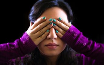 A woman with long dark hair covers her eyes with her hands. She has turquoise nail polish and wears a purple sweater with button details on the sleeves. The background is black.